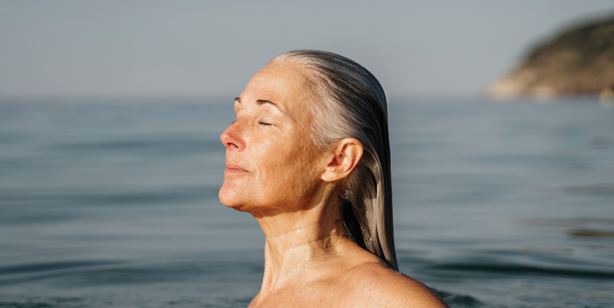 elderly woman standing in the clean sea water with serene expression and eyes closed, enjoying the sunlight on her face