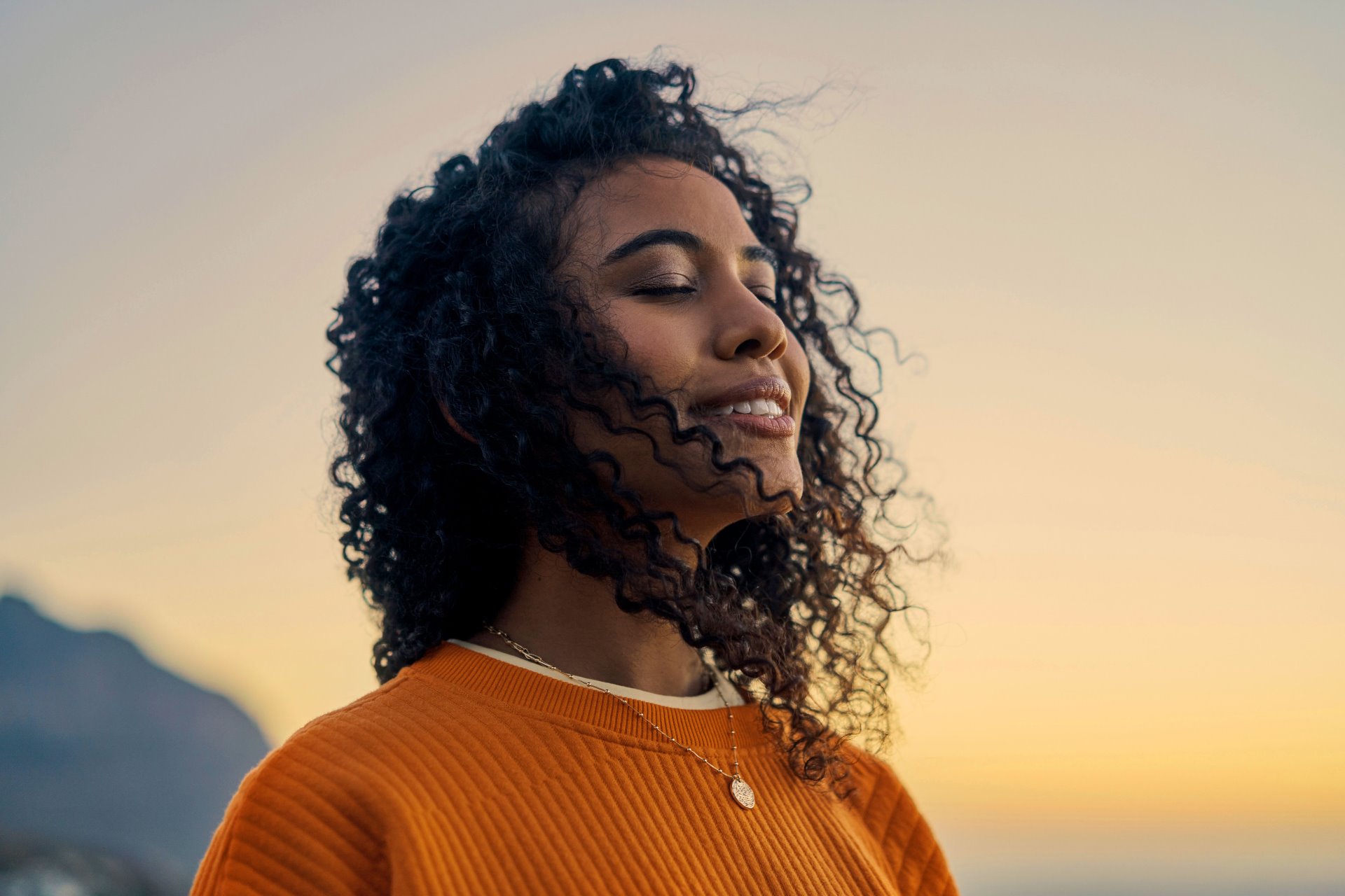 a young woman with curly hair, eyes closed, smiling gently as she enjoys the pleasant cool breeze at sunset